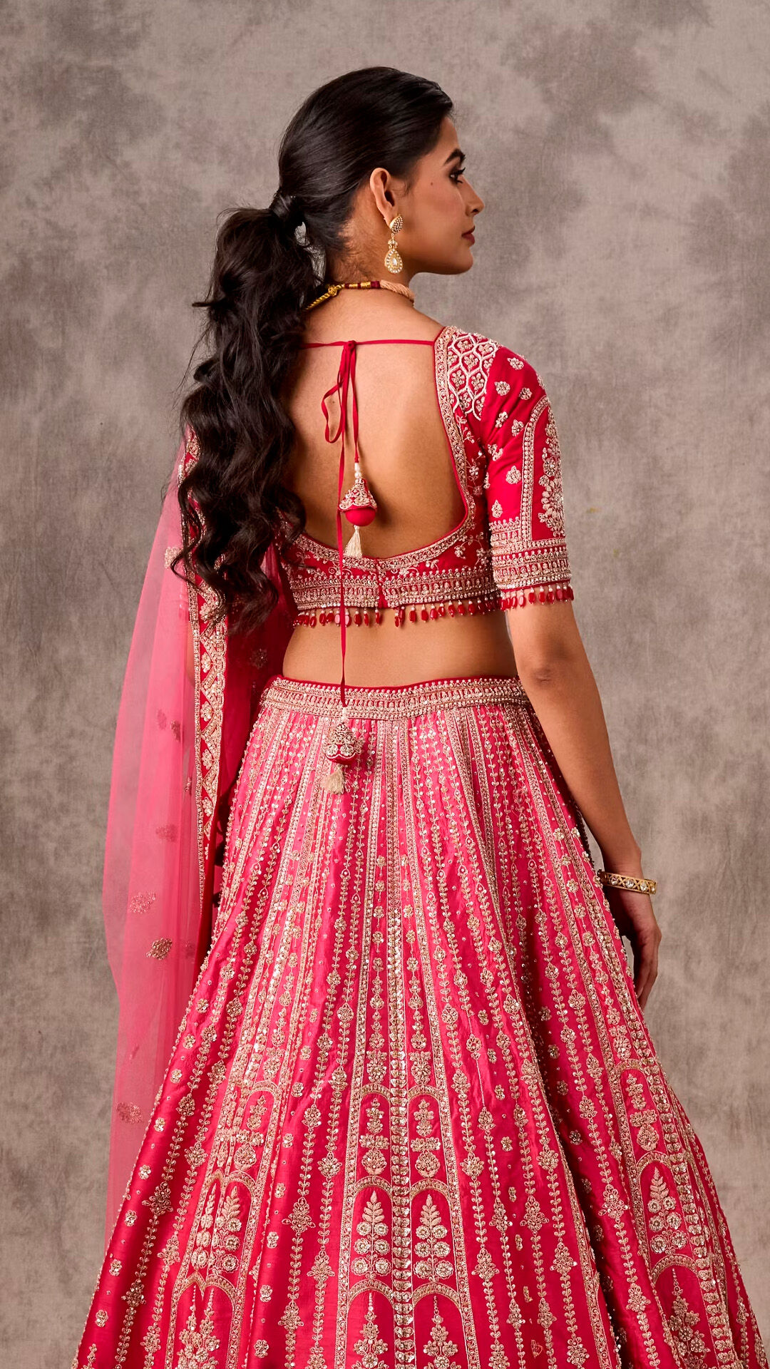 Bride posing in a hot pink lehenga, showing the elaborate backless blouse, tassel detail, and a voluminous wavy ponytail hairstyle.